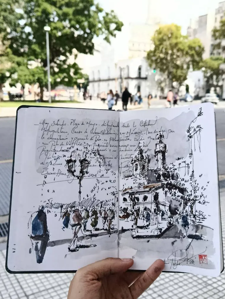 El mismo sketchbook abierto con Plaza de Mayo y la Catedral en tinta y aguada, notas manuscritas y sello rojo, frente al Cabildo
