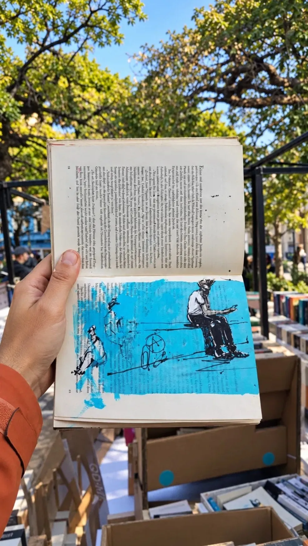Libro antiguo intervenido con una figura en tinta sobre una mancha azul, sostenido en la mano entre los puestos de una feria de libros