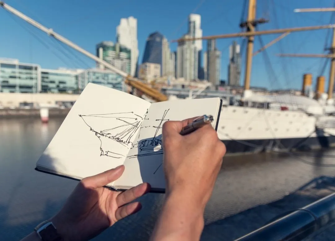 Mano trazando en tinta un velero en un sketchbook, frente a la Fragata Sarmiento y las torres de Puerto Madero