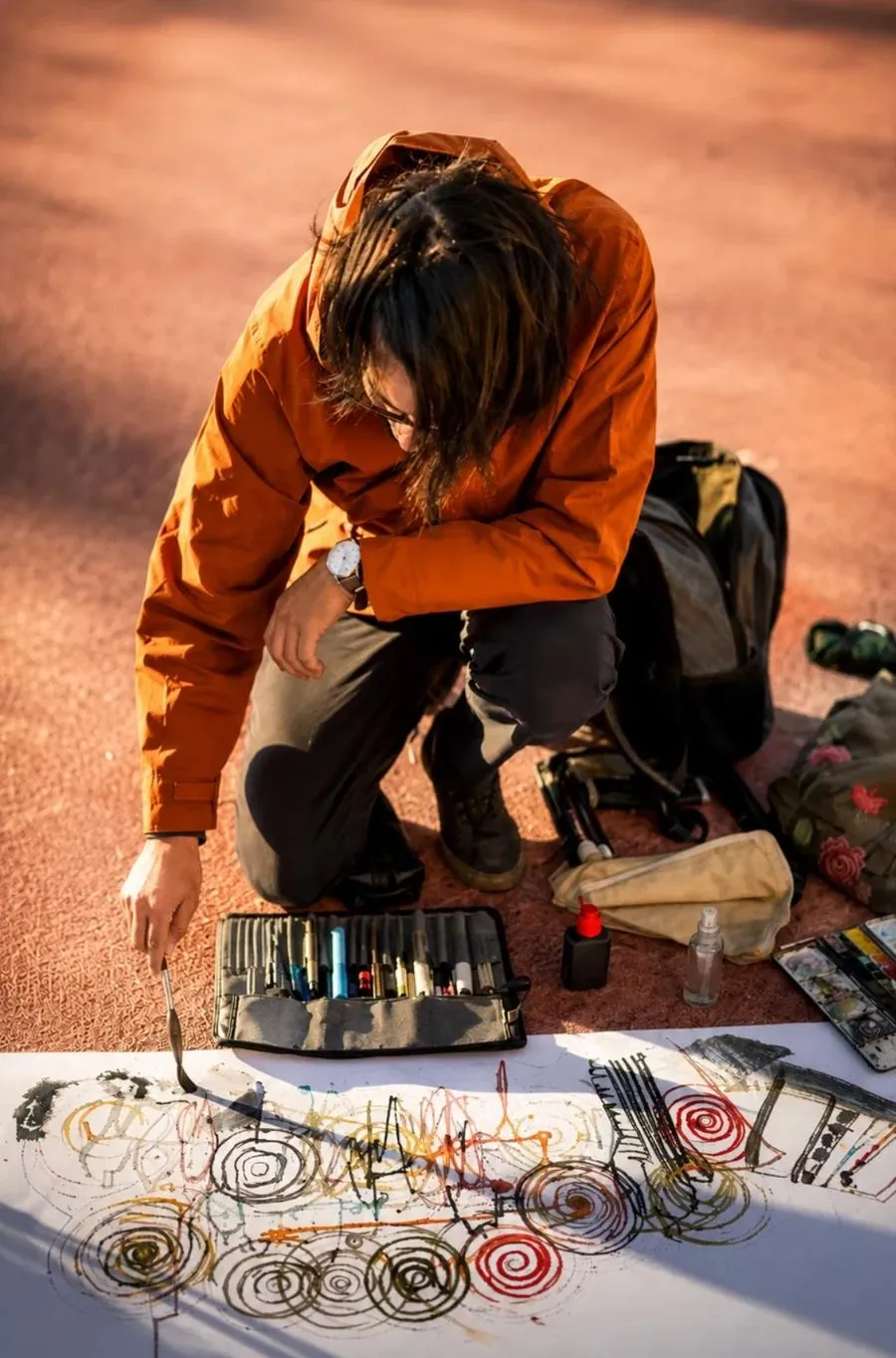 William, con campera naranja, arrodillado en el suelo dibujando espirales de tinta y color sobre un papel blanco de gran formato, con sus materiales alrededor