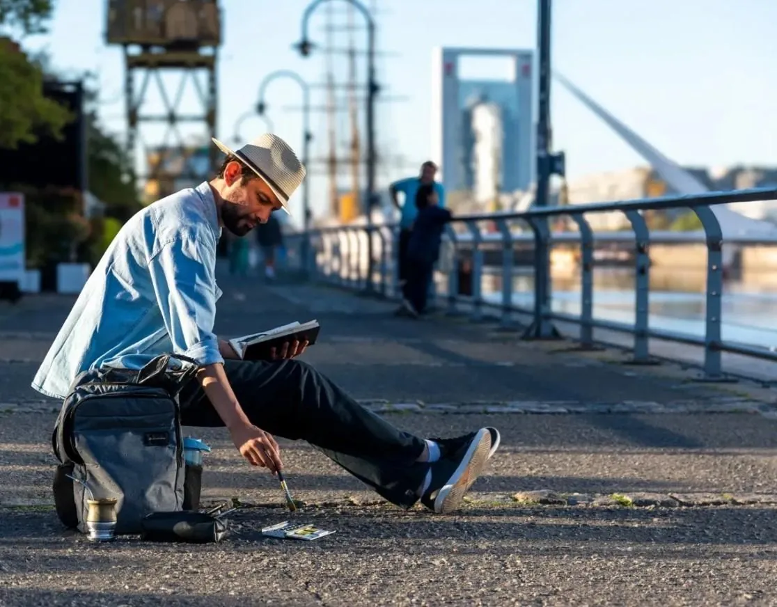 William dibujando sentado en el suelo de Puerto Madero, con su mochila, el mate y el Puente de la Mujer detrás