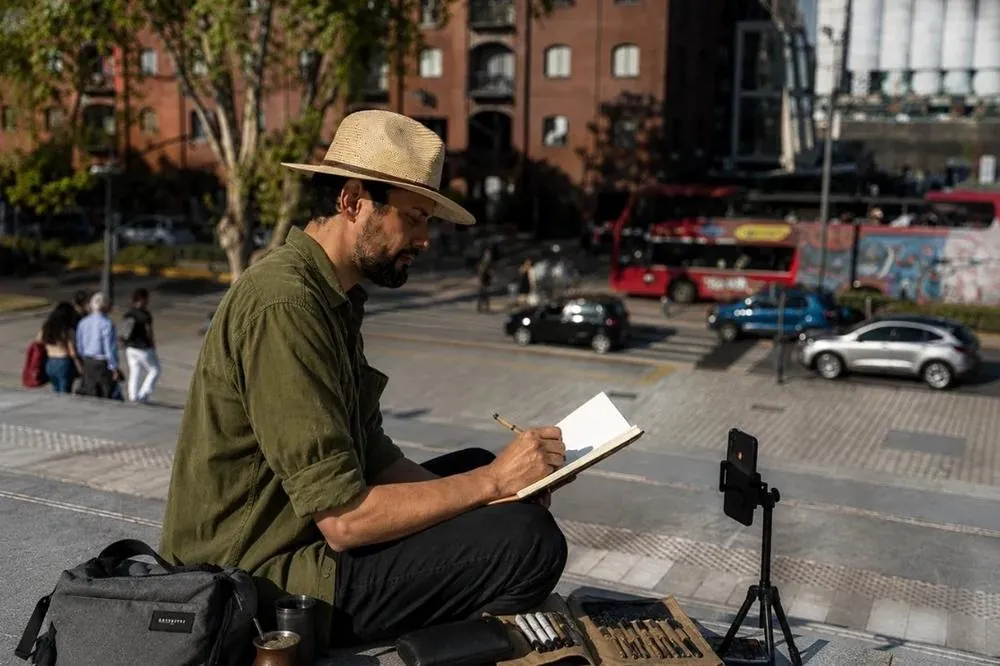 William dibujando sentado en una escalinata, con sombrero, el estuche de plumas abierto y un trípode con el celular grabando